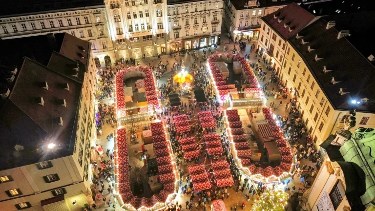 Le marché de Noël de la place principale de Bratislava vu du ciel de nuit, ses étals rouges et blancs remplissant la place autour d'un carrousel illuminé.