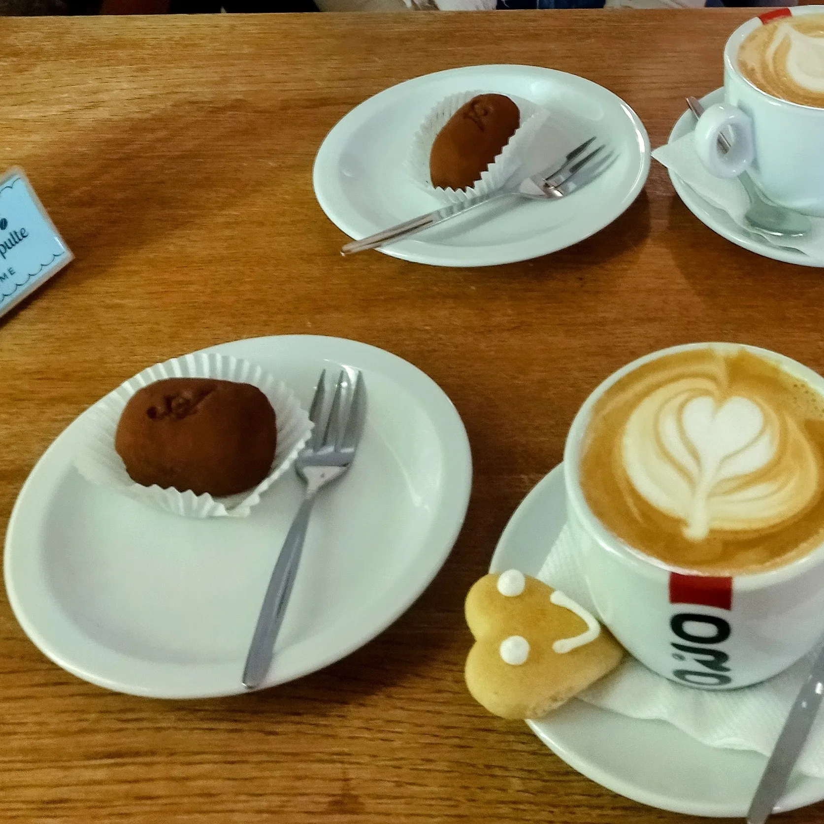 A cozy café setting featuring a cappuccino with heart-shaped latte art, accompanied by a smiling heart-shaped cookie. On the table, there are two servings of Kremnický Krumpel, a traditional handmade chocolate delicacy from Kremnica, Slovakia, with a soft, cocoa-dusted exterior. The warm wooden table and elegant white plates add to the inviting atmosphere
