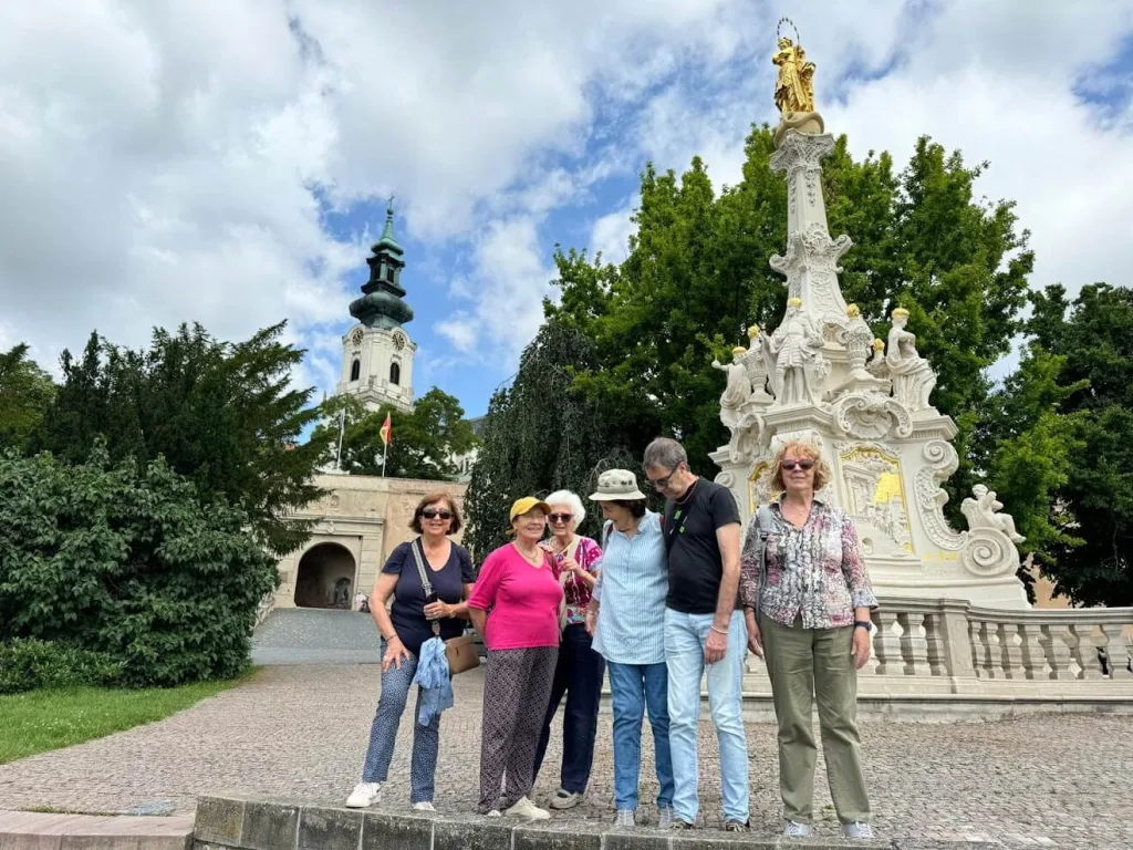 A group of tourists standing on a cobblestone plaza in Nitra during Central European Itinerary, Slovakia, next to an ornate white Baroque monument with gold accents topped with a religious statue. In the background is a church tower with a green spire and an archway entrance to what appears to be a castle or historic complex, all under a partly cloudy sky.