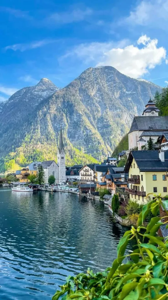 A breathtaking view of Hallstatt, Austria, with its picturesque village nestled between a clear lake and towering Alpine mountains. The scene features charming traditional Austrian buildings including a church with a tall spire, colorful houses along the waterfront, and small boats docked at the shore. The dramatic mountain backdrop rises steeply behind the village with some trees visible on the lower slopes, all under a bright blue sky with scattered clouds.