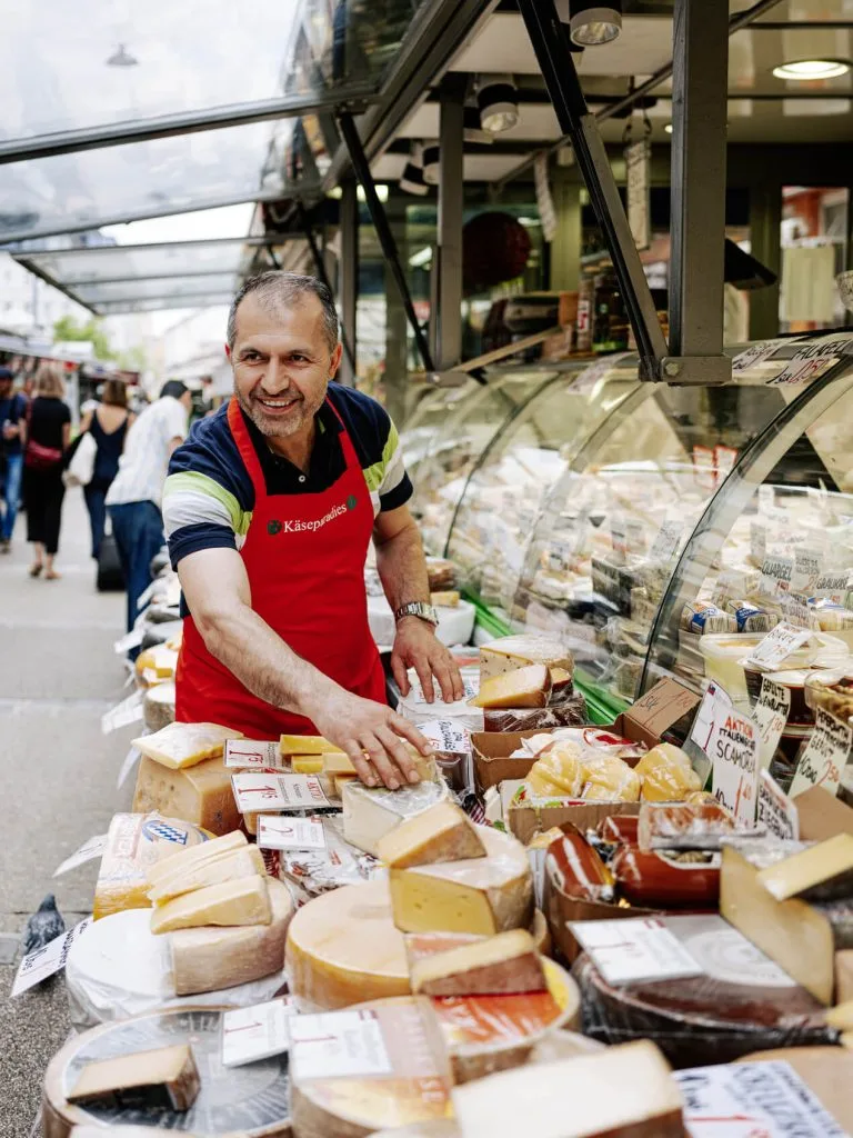 Un fromager souriant sur un étal du Naschmarkt à Vienne, derrière un comptoir de meules entières et de portions.