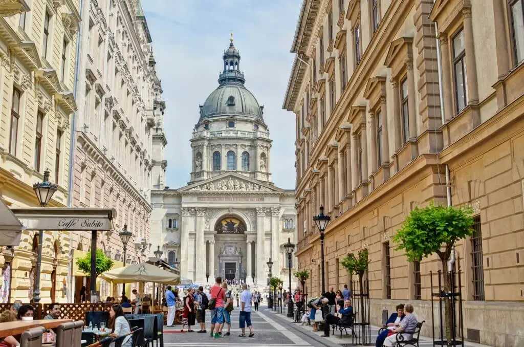 St Stephen's Basilica in Budapest at the end of a pedestrian street lined with café terraces and clipped trees.