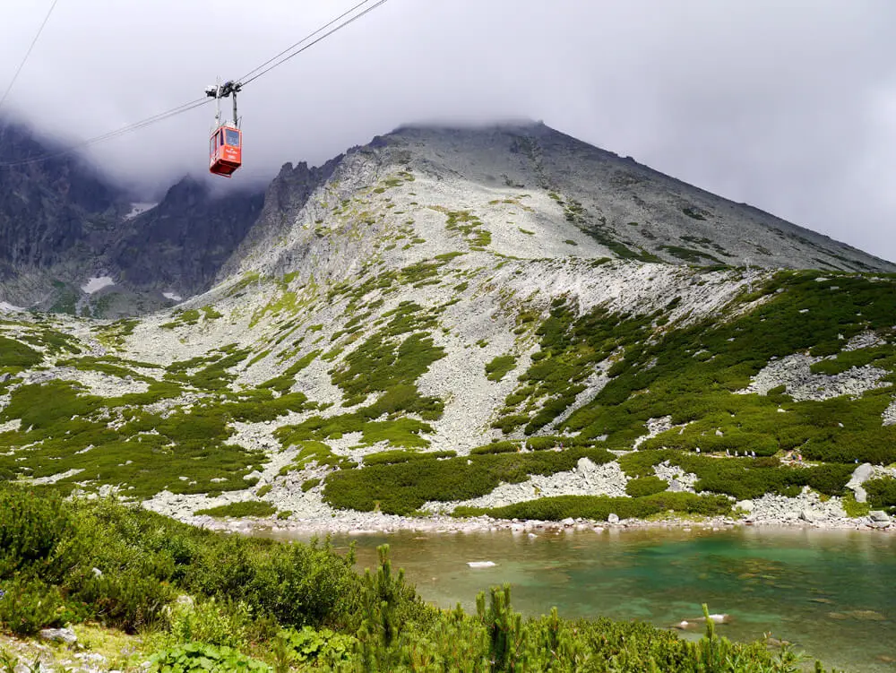 A cable car crossing above a green mountain tarn in the High Tatras, scree slopes and cloud behind.