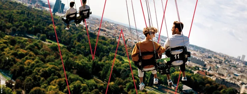 Riders on a high chain-swing ride at the Prater, swinging out above the trees with the rooftops of Vienna below.