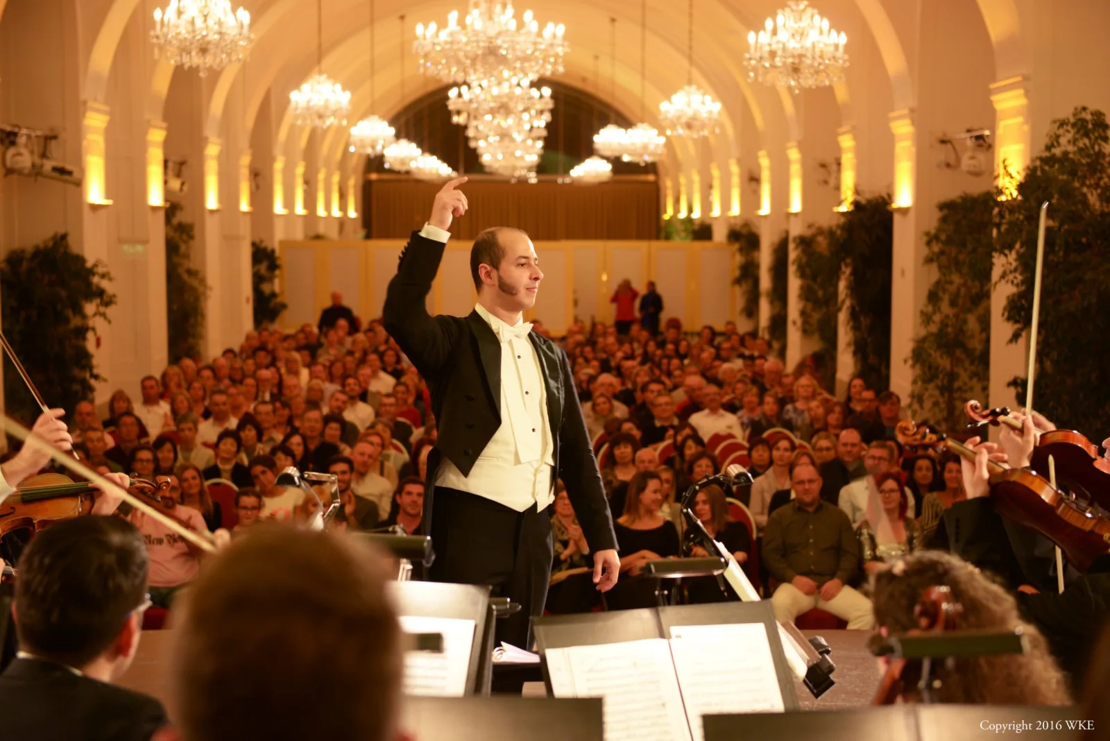 A conductor and orchestra performing to a full audience in a chandeliered hall.