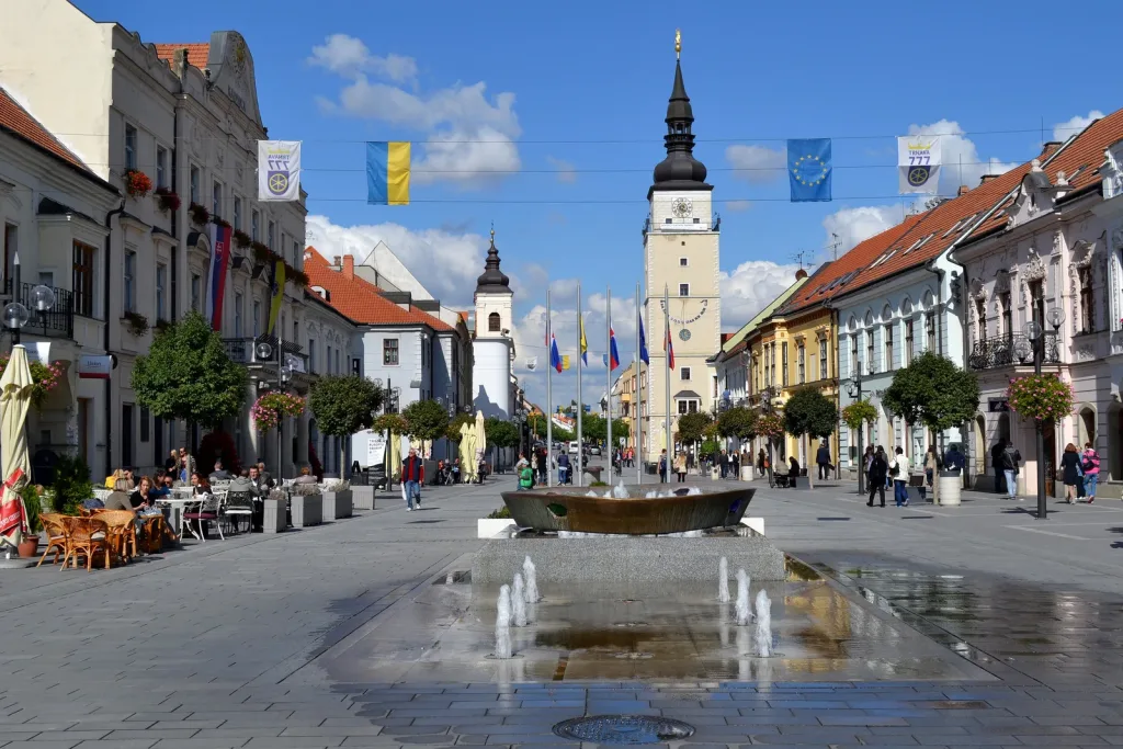 Trnava's main square looking towards the white City Tower, with café terraces, flags strung across the street and a fountain in the paving.