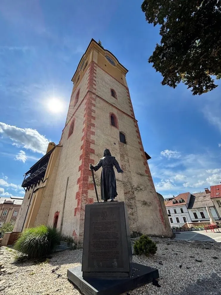 A medieval stone tower in the castle precinct of Banská Bystrica, with a bronze statue of King Béla IV standing before it.