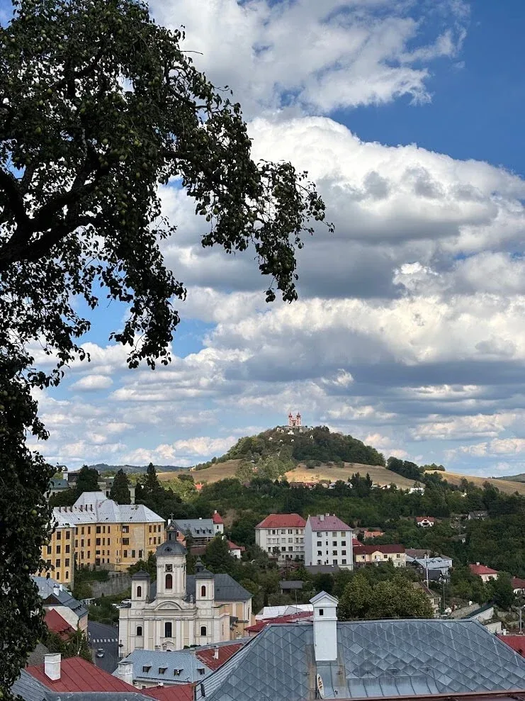 A view over the roofs of Banská Štiavnica to the Calvary on its wooded hill, with a baroque church in the foreground.