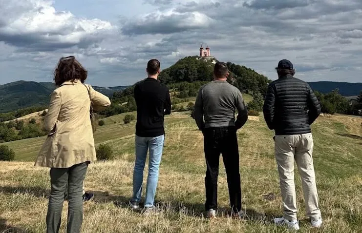 Four visitors standing on a meadow looking across to the Calvary above Banská Štiavnica, its chapels rising on the wooded hilltop under a wide sky.