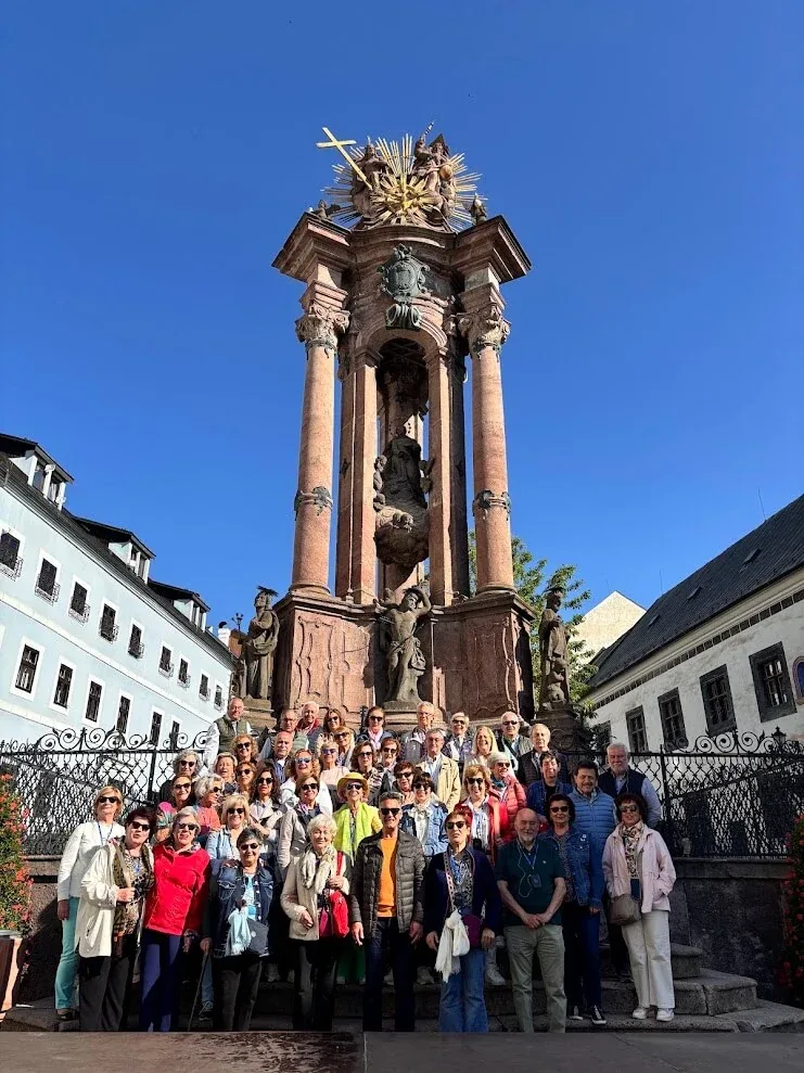 A large tour group gathered on the steps of the Holy Trinity column in Banská Štiavnica on a clear morning.