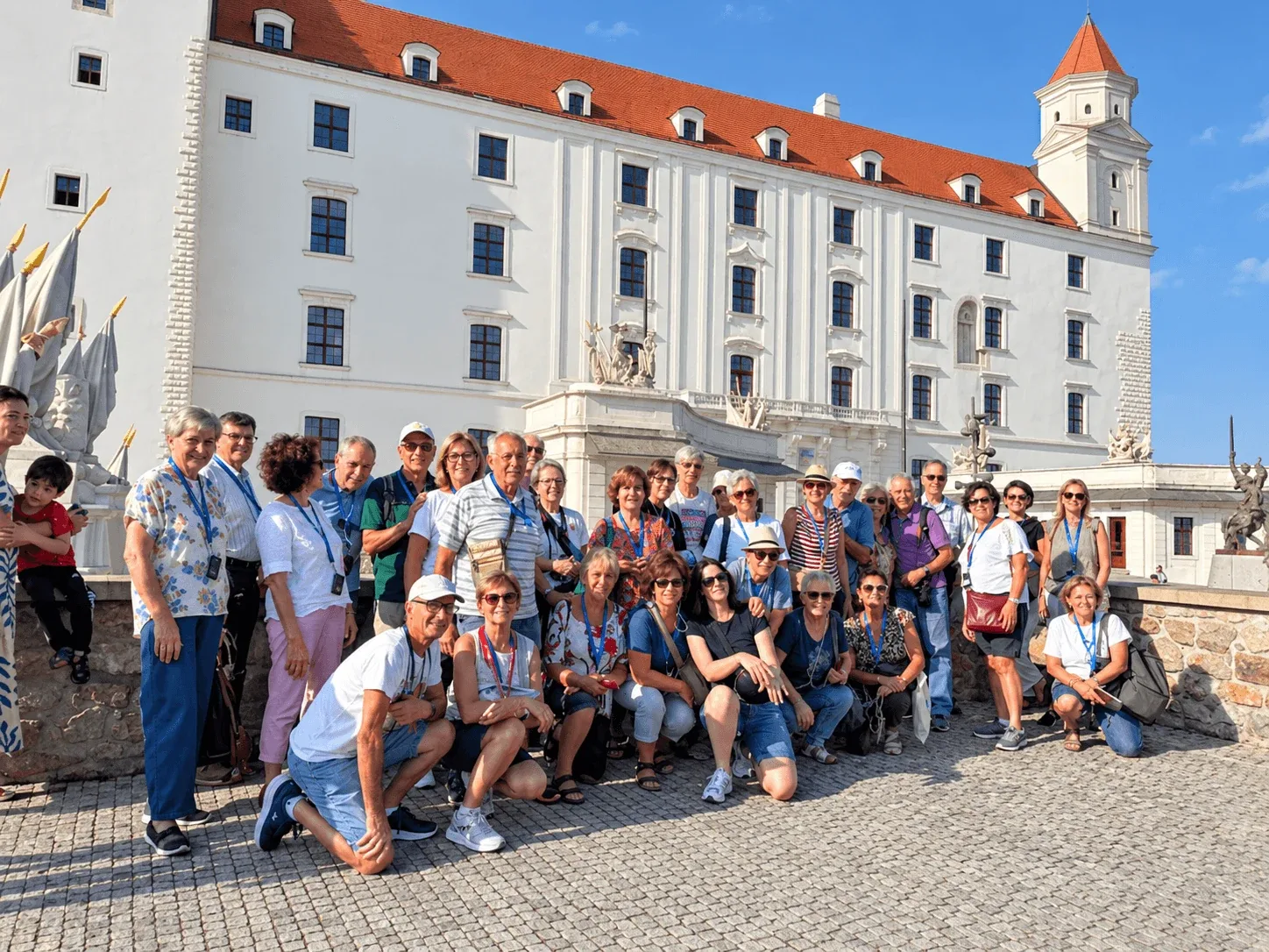 A tour group wearing audio-guide lanyards photographed in front of Bratislava Castle, its white facade and red roof rising behind them.