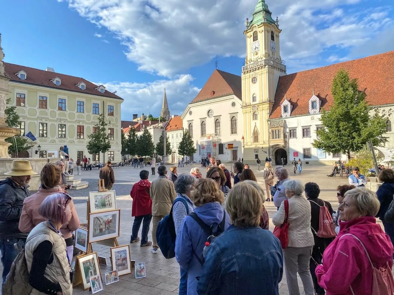 Un groupe de touristes sur la place principale de Bratislava, avec des aquarelles à vendre sur des chevalets et la tour de l'ancien hôtel de ville derrière.