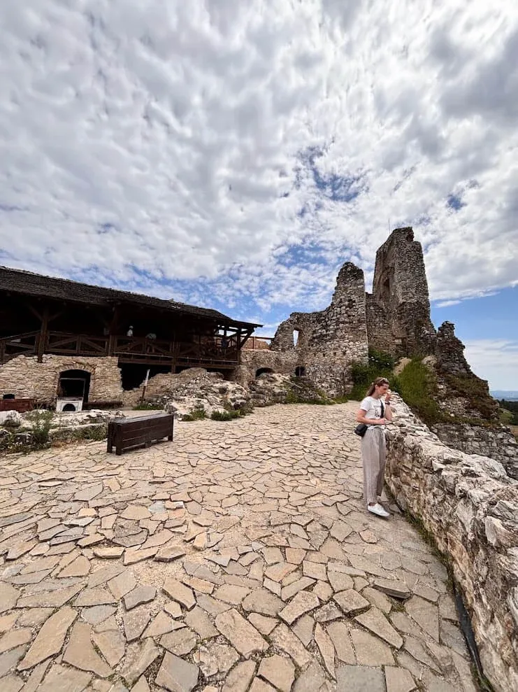 A visitor on the stone-paved courtyard of the ruined Čachtice Castle, its broken towers standing against a cloudy sky.