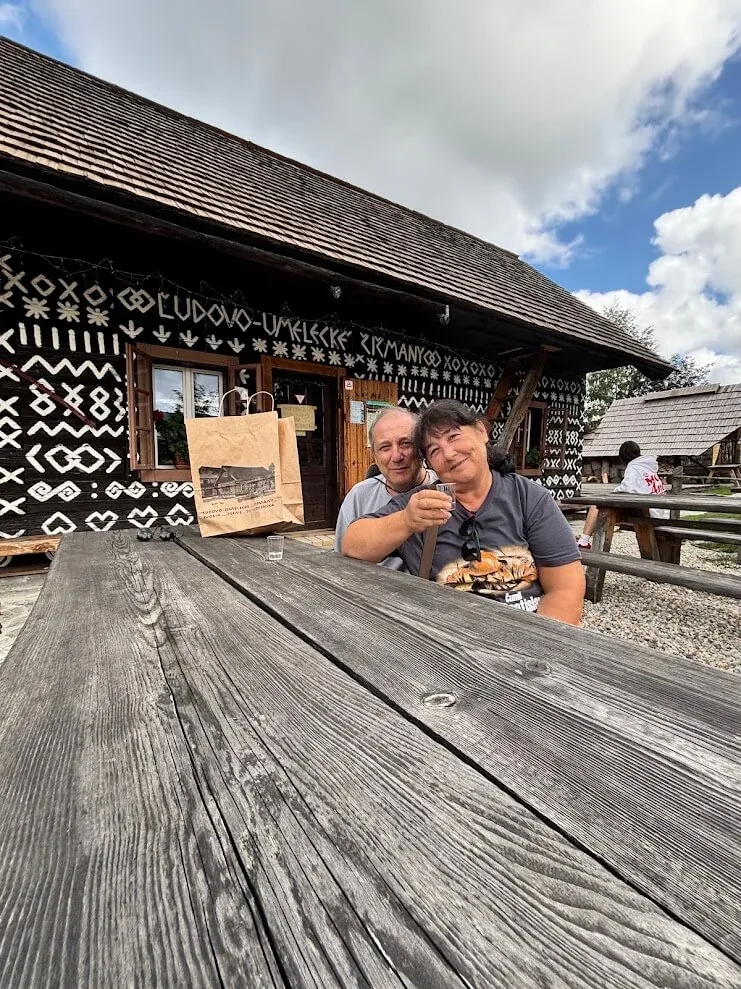 Two visitors at a wooden table outside a painted folk-art house in Čičmany, raising a glass in the sunshine.