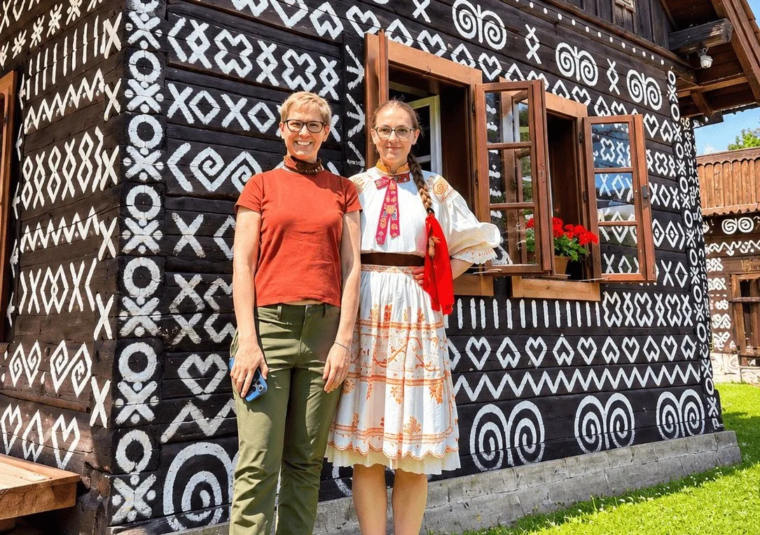 Un visitante y una mujer con traje folclórico tradicional eslovaco frente a una casa de madera pintada en Čičmany.