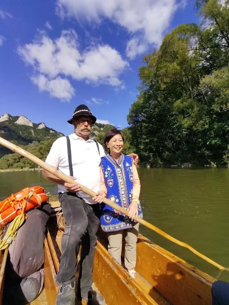 A raftsman in traditional dress with a guest aboard a traditional wooden raft on the Dunajec River, the wooded hills of the Pieniny rising behind them.