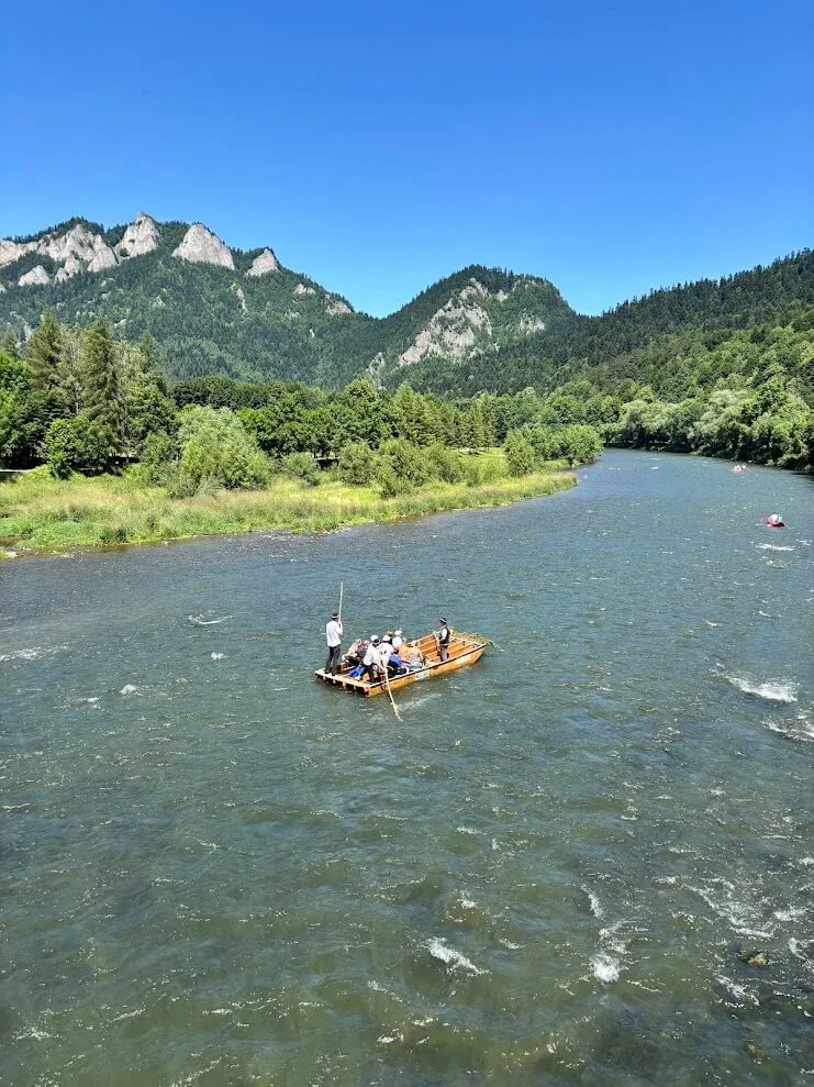 A wooden raft carrying passengers down the Dunajec River, with the rocky peaks of the Pieniny rising behind the wooded banks.