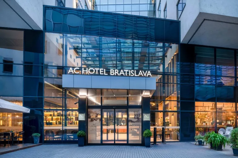 The entrance of AC Hotel by Marriott Bratislava Old Town at dusk, its lobby and lounge lit up behind the glass front.
