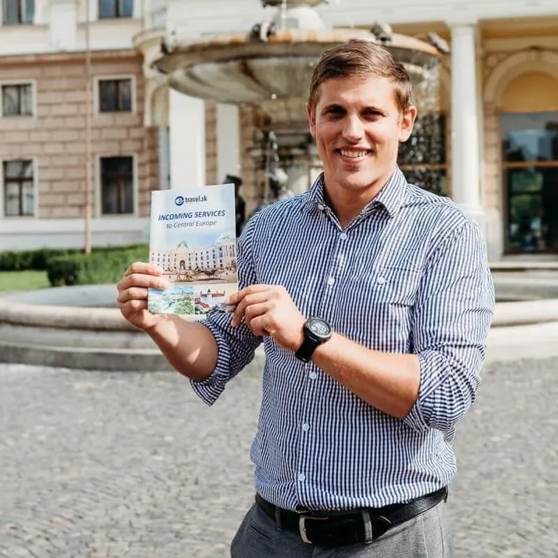 Andrej Stanik, VIP-Guide bei E-TRAVEL.SK, hält neben einem Brunnen die Broschüre „Incoming Services to Central Europe“ des Unternehmens.