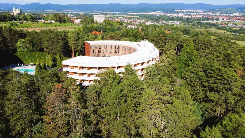 An aerial view of a curved spa hotel among the pine woods at Bojnice, with Bojnice Castle in the distance.