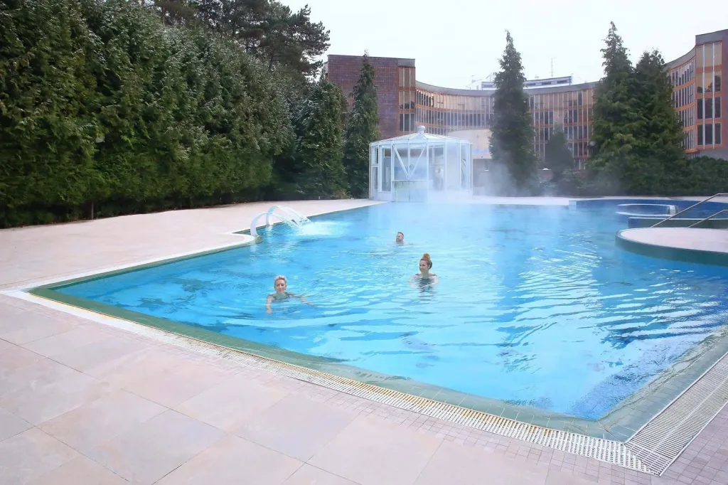 Guests swimming in a steaming outdoor thermal pool at the Bojnice spa, with the curved spa hotel behind.