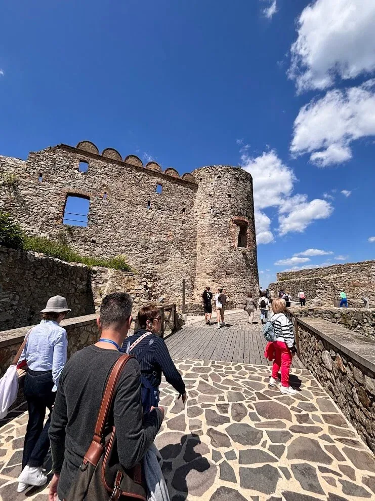 Visitors walking up the stone path through the ruined walls and round tower of Devin Castle near Bratislava under a bright summer sky.