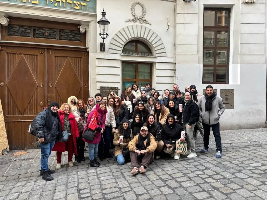A large tour group posing together on a cobbled street in Vienna's old town, in front of a tall wooden gateway with a Hebrew inscription above it.