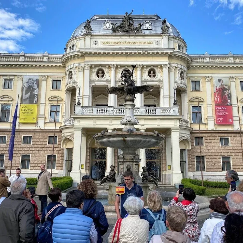 A tour group standing in front of the historic Slovak National Theatre in Bratislava, with the Ganymede Fountain playing between them and the building.