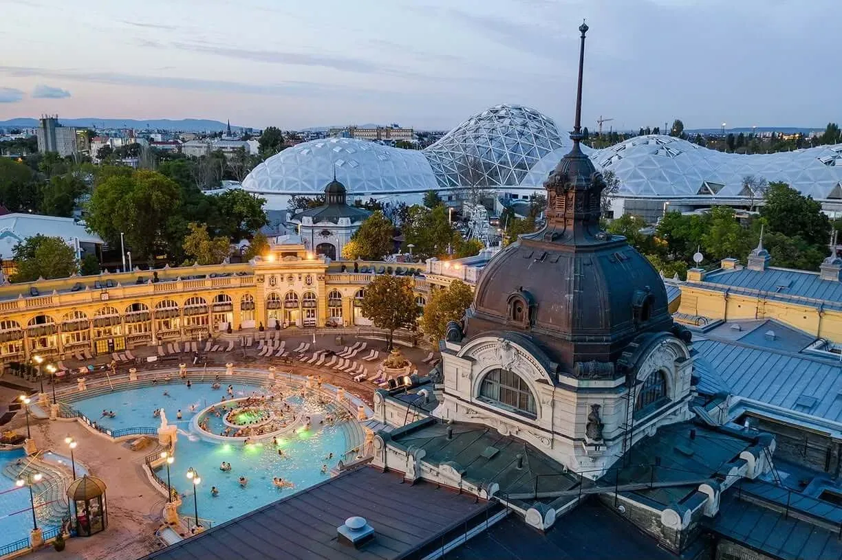 Las piscinas termales exteriores del Balneario Széchenyi de Budapest al anochecer, rodeadas por las arcadas neobarrocas amarillas del complejo termal.