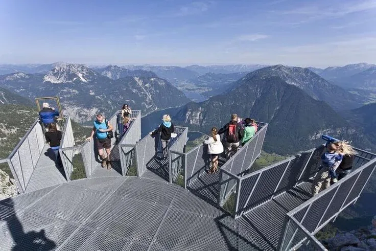 Visitors walking out onto the Five Fingers viewing platforms high in Austria's Dachstein mountains, with the lake and surrounding peaks far below.