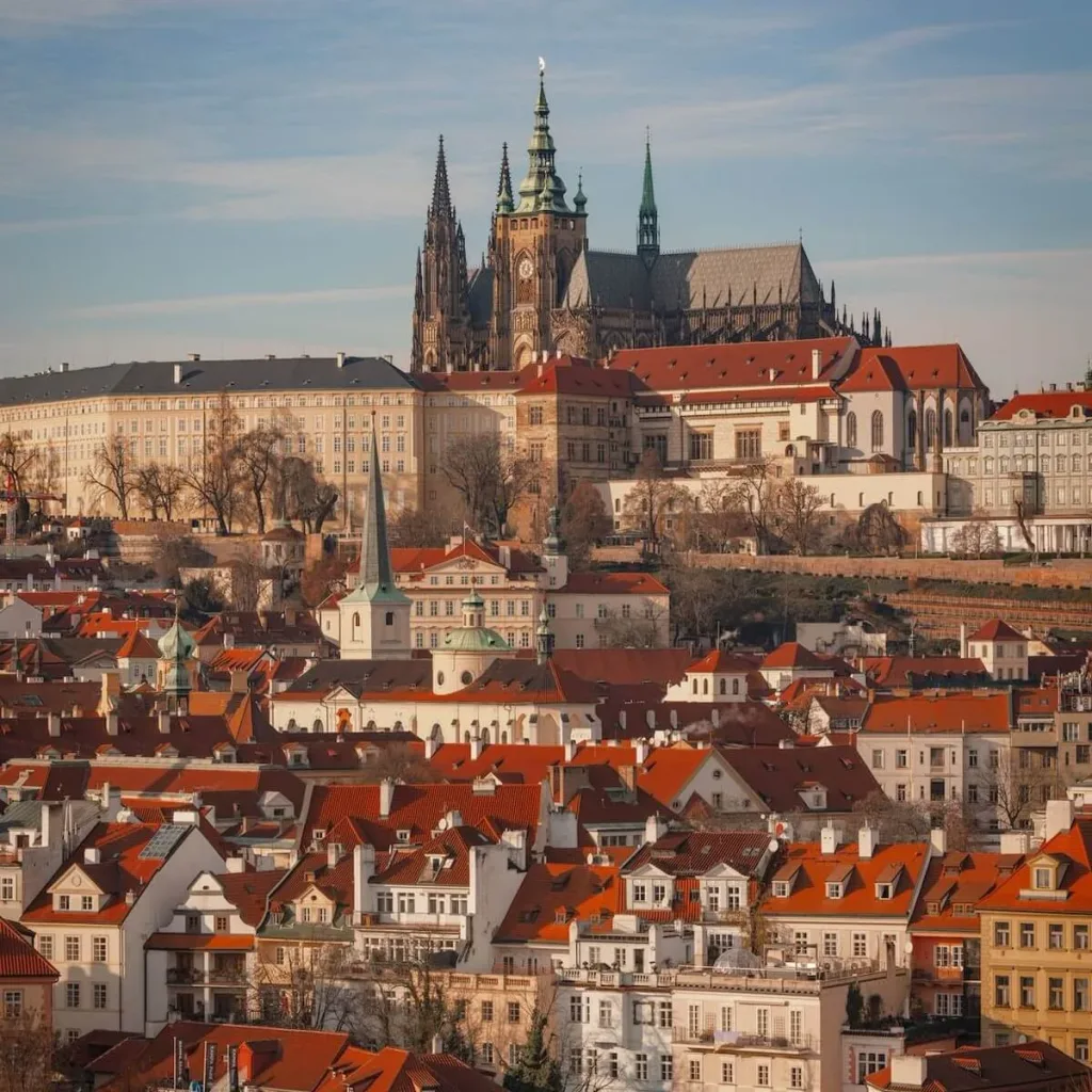 Prague Castle and St Vitus Cathedral rising above the red rooftops of the Lesser Town in golden evening light.