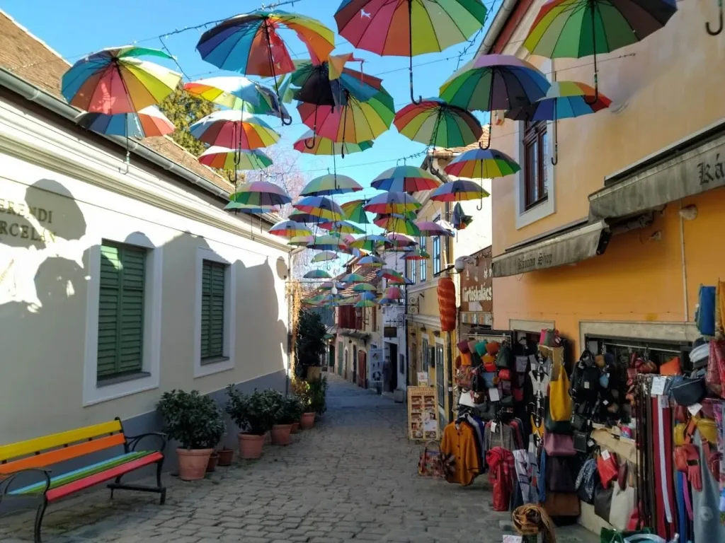A cobbled lane in Szentendre, Hungary, hung with rows of brightly coloured umbrellas above pastel houses and souvenir shops.