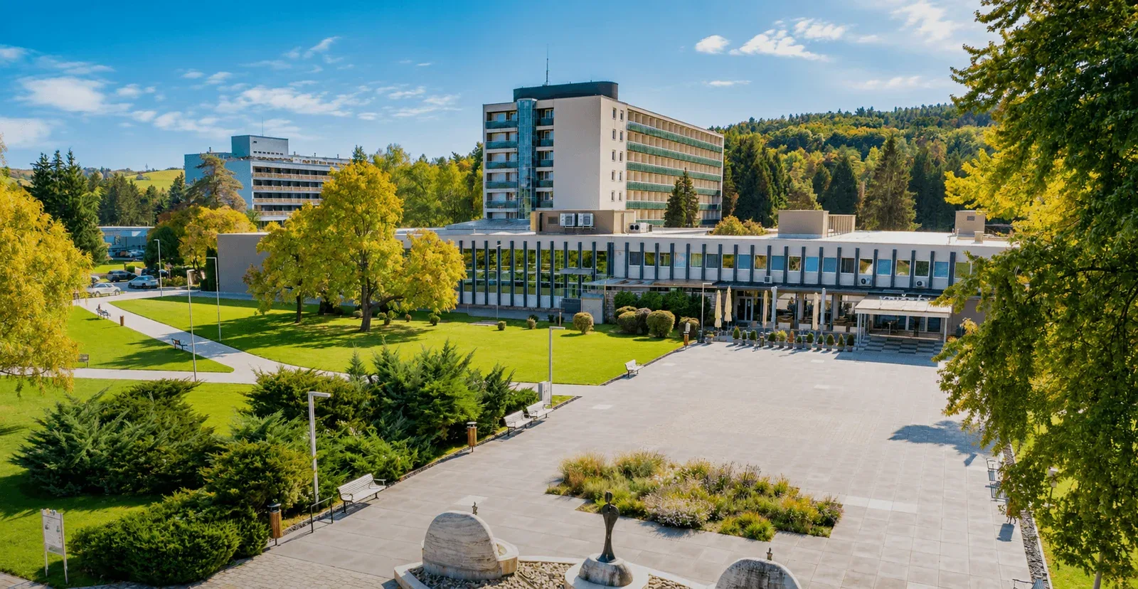 The spa promenade in Dudince in autumn sunshine, with the spa hotels behind the lawns and a sculpture fountain in the foreground.