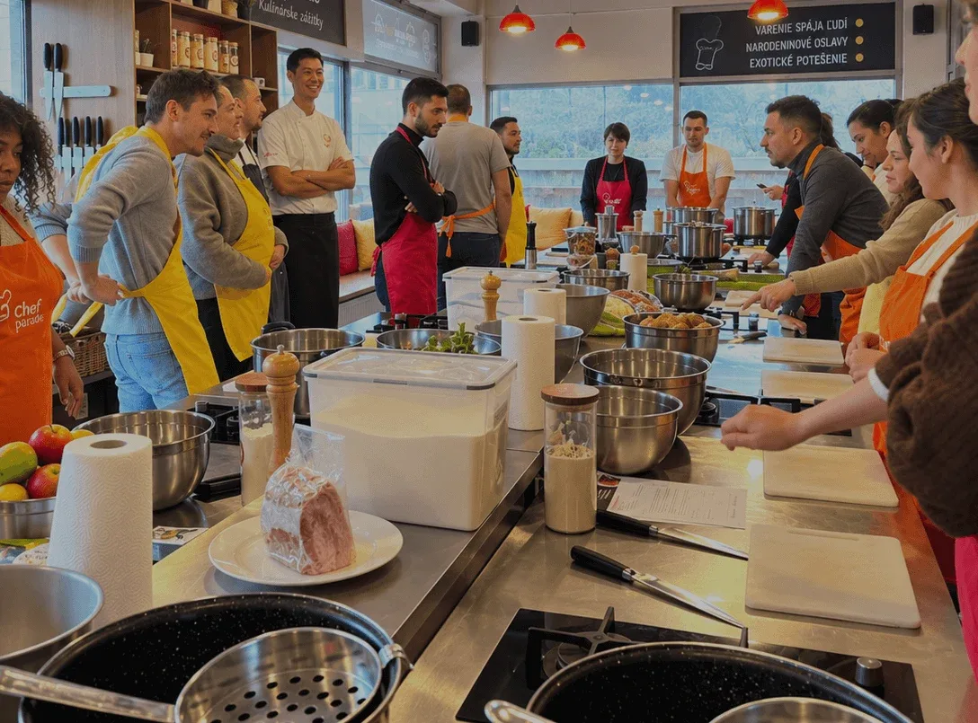 Un corso di cucina di gruppo alla scuola di cucina Chefparade, con i partecipanti in grembiule riuniti attorno ai piani di lavoro della cucina e uno chef che guida la sessione.