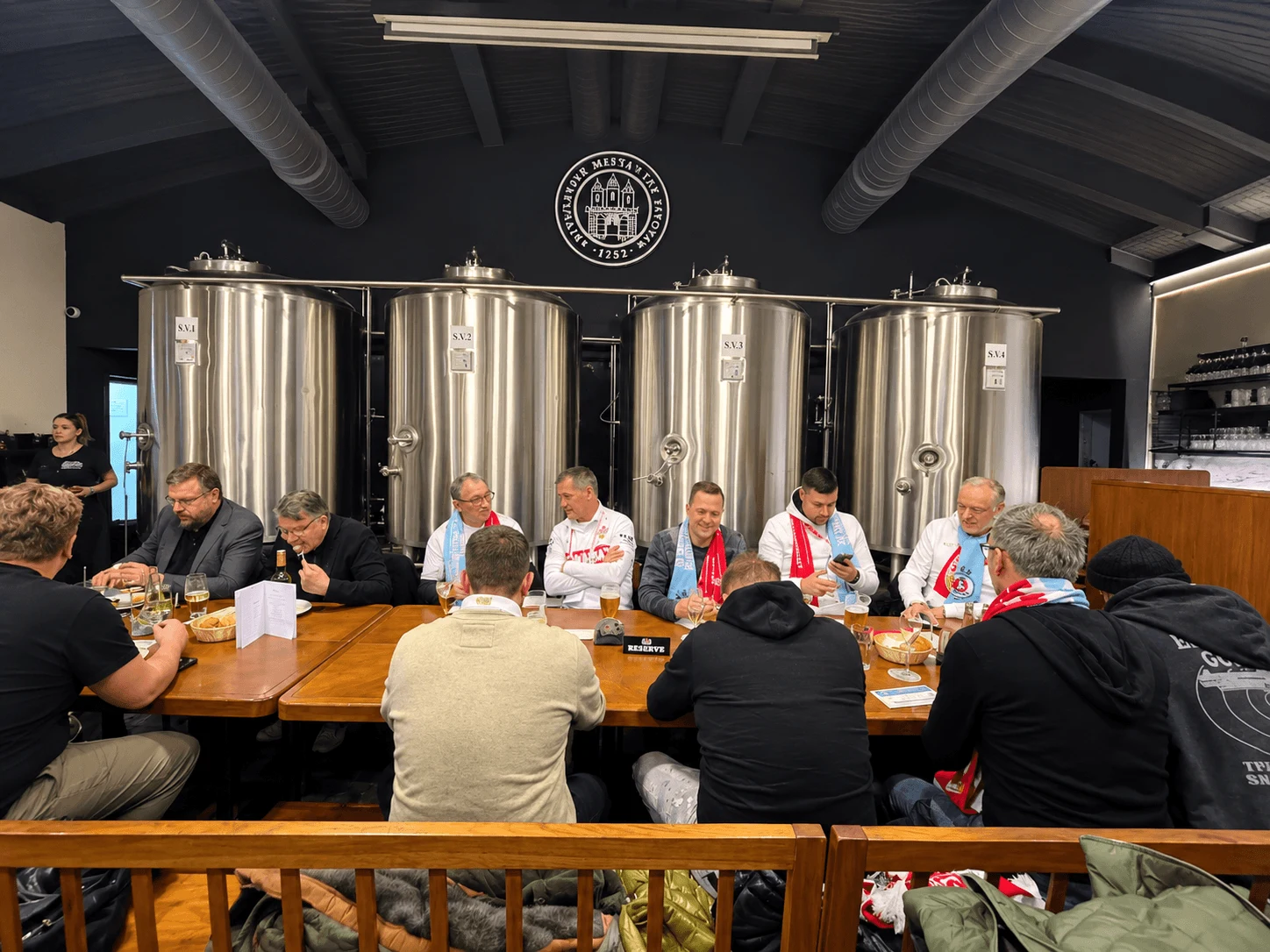 Un groupe à une longue table dans la salle de brassage de la brasserie des Bourgeois de Bratislava (Bratislavský meštiansky pivovar), avec ses cuves de brassage en acier derrière eux.