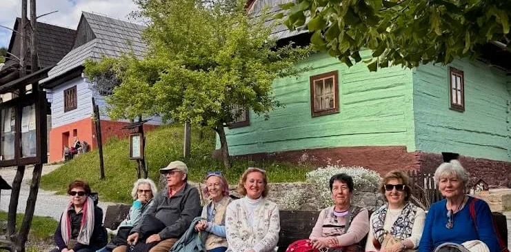Un gruppo di turisti sorridenti seduto su una panchina davanti a case di tronchi dipinte di blu e verde in un villaggio slovacco.