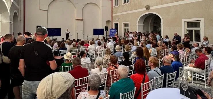 Un public assis dans une cour historique pour une conférence en plein air, avec un orateur sur scène entre deux écrans.