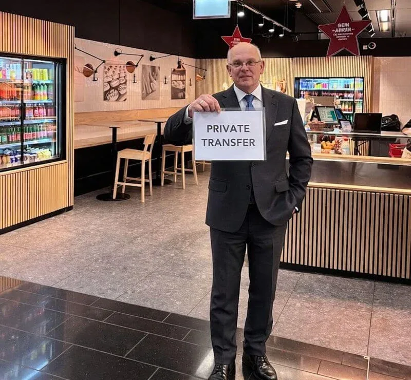 Un conductor de traje sosteniendo un cartel de «Private transfer» en una sala de llegadas, esperando a sus huéspedes.