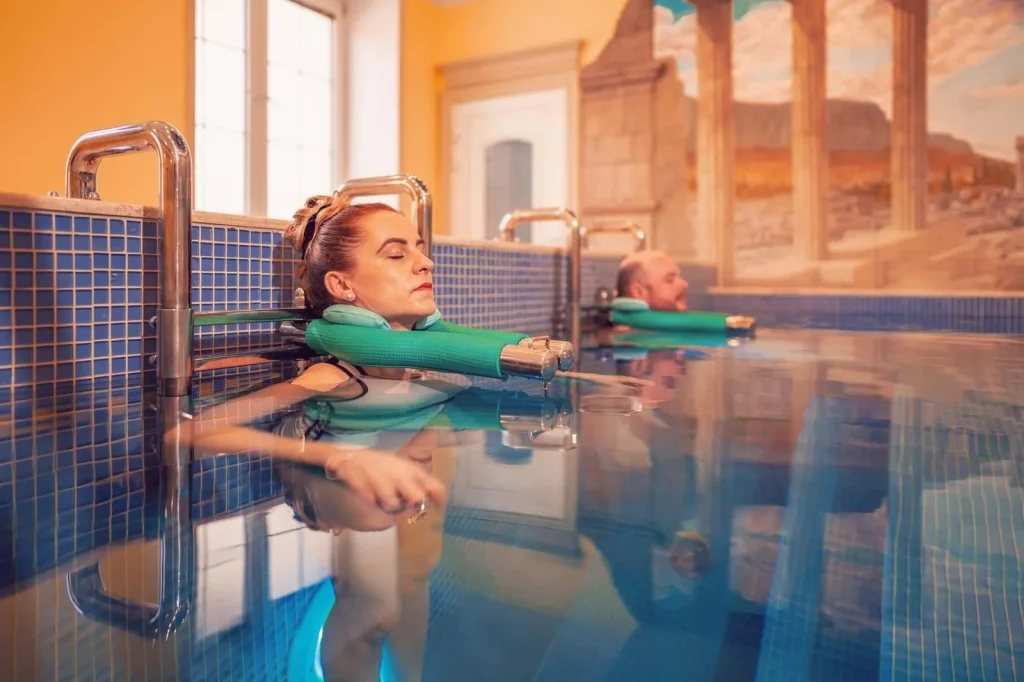 Guests floating with neck supports in a thermal pool at the Aphrodite spa in Rajecké Teplice, beneath a mural of classical ruins.