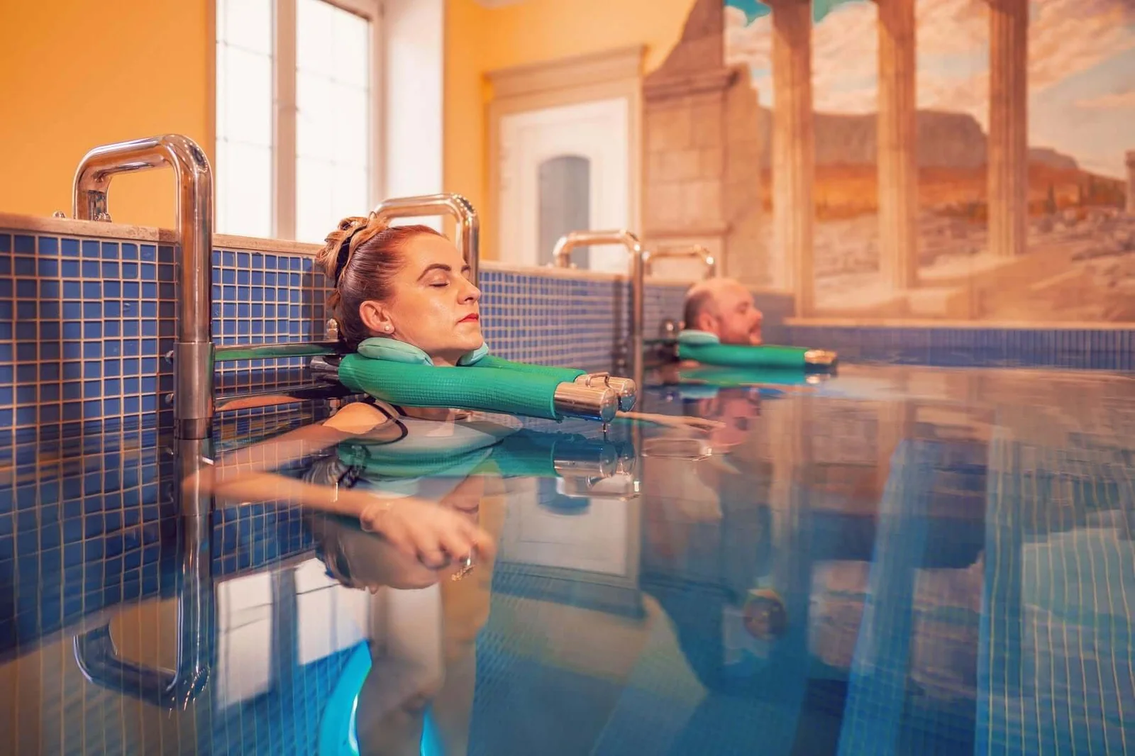 Guests floating with neck supports in a thermal pool at the Aphrodite spa in Rajecké Teplice, beneath a mural of classical ruins.
