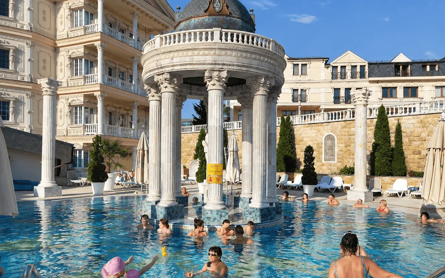 Открытый термальный бассейн курорта Aphrodite в Раецке-Теплице, купающиеся вокруг ротонды с колоннадой, позади курортный отель.