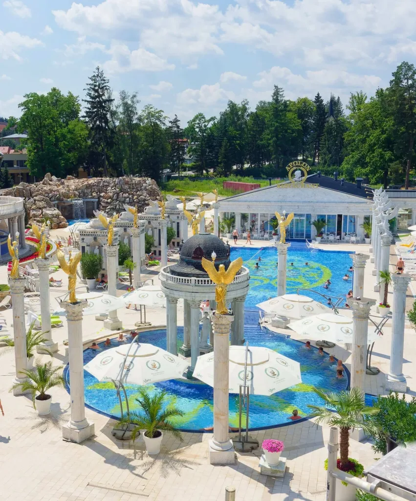Bathers in the outdoor thermal pools at the Aphrodite spa in Rajecké Teplice on a summer day, among white columns, golden angel statues and parasols.