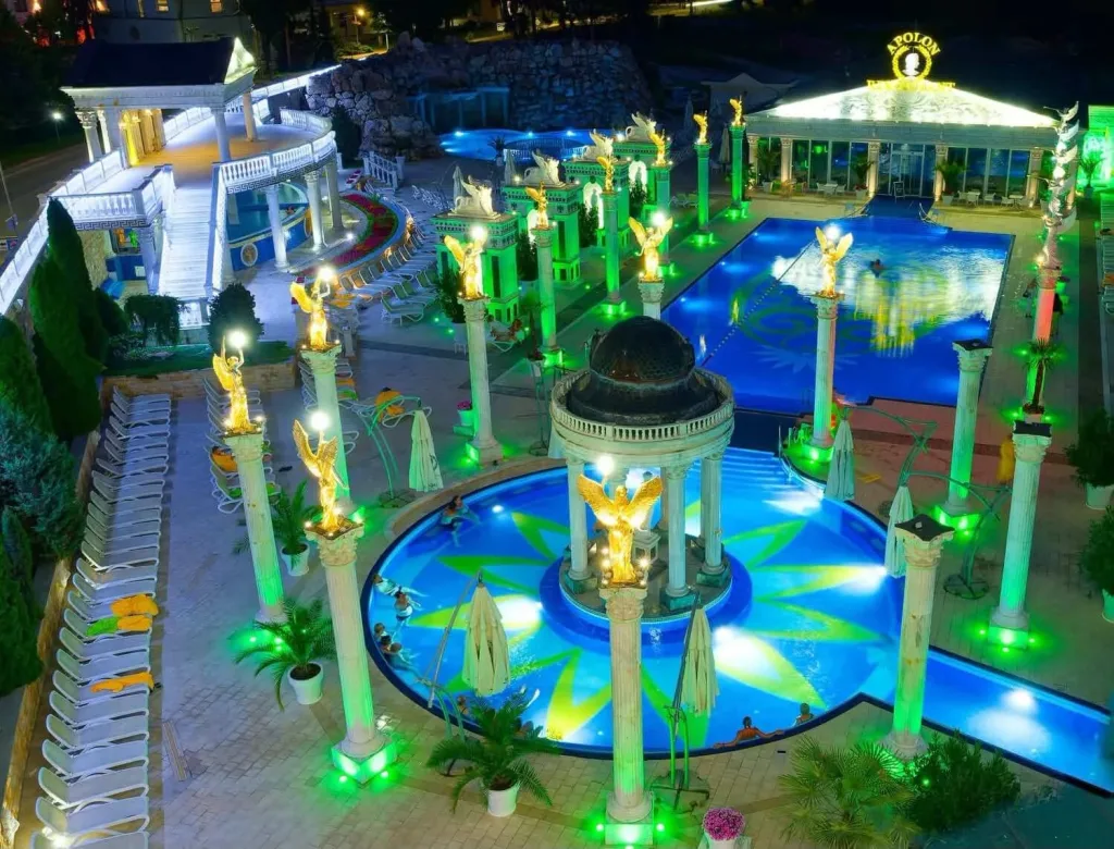 The outdoor thermal pools at the Aphrodite spa in Rajecké Teplice lit up at night, with golden angel statues on white columns around the rotunda.