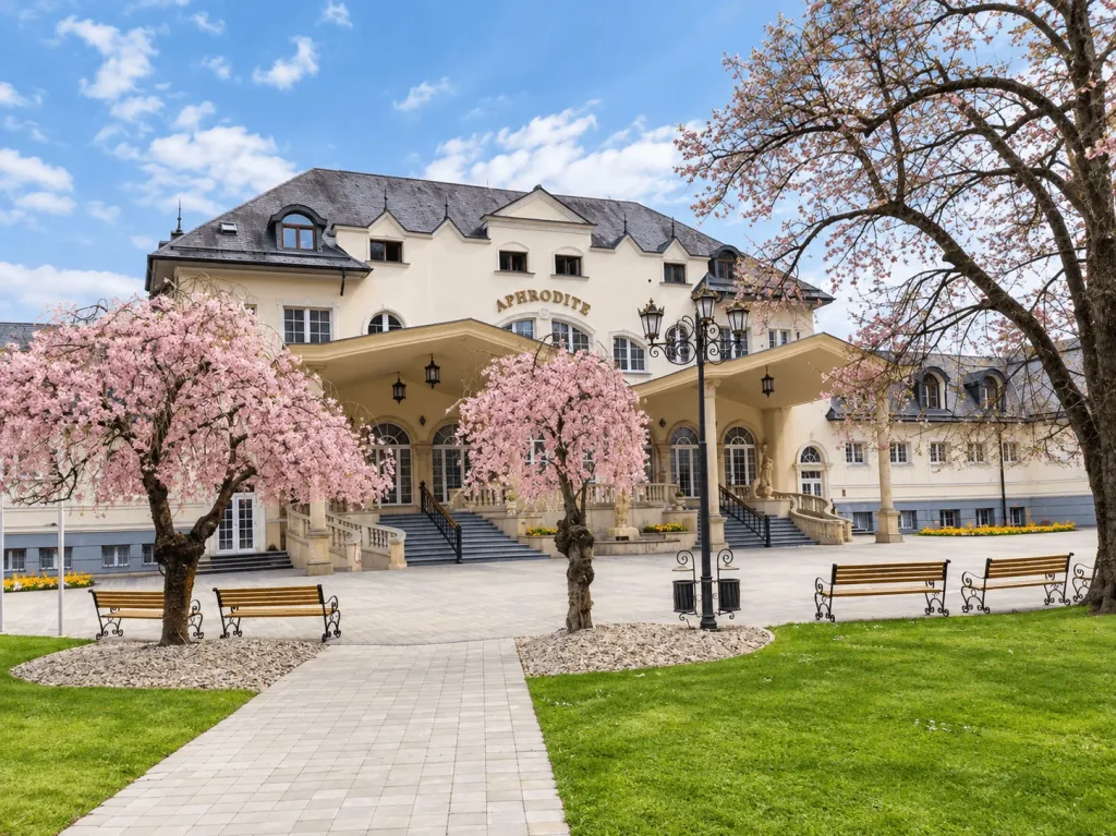 The Aphrodite spa building in Rajecké Teplice, with pink cherry trees in blossom and benches in the spa park in front.