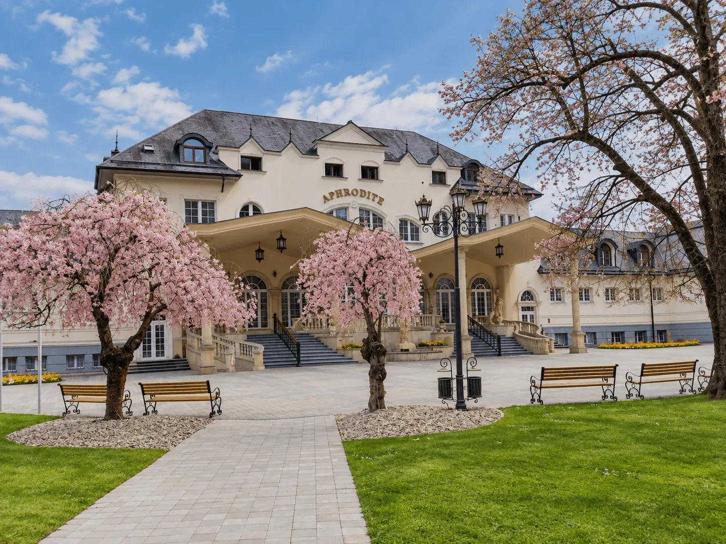 The Aphrodite spa building in Rajecké Teplice, with pink cherry trees in blossom and benches in the spa park in front.