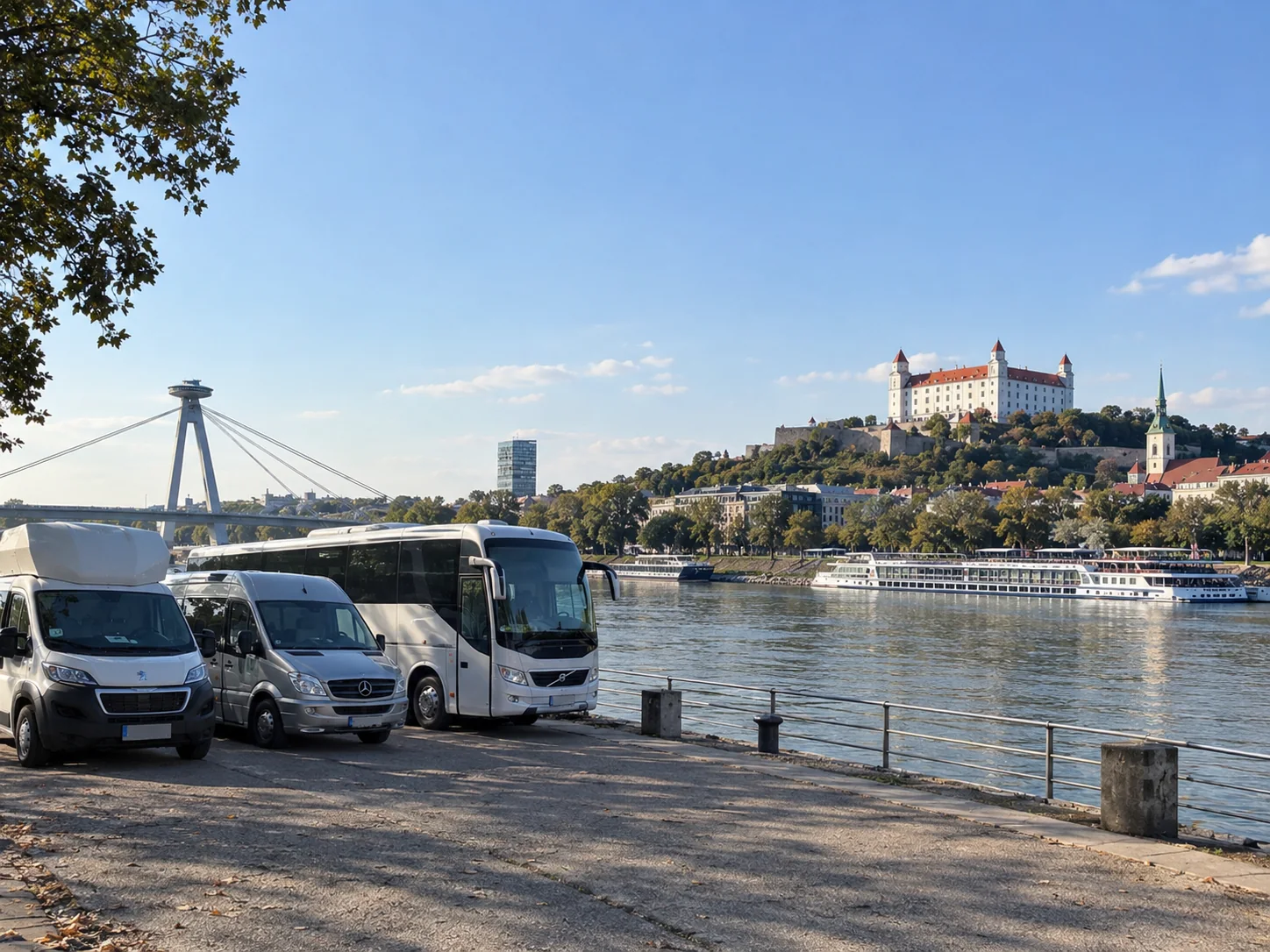 Ein Reisebus, ein Minibus und ein Van stehen an der Donaupromenade in Bratislava, unterhalb der Burg Bratislava liegen Flusskreuzfahrtschiffe vor Anker, in der Ferne der Most SNP (die UFO-Brücke).