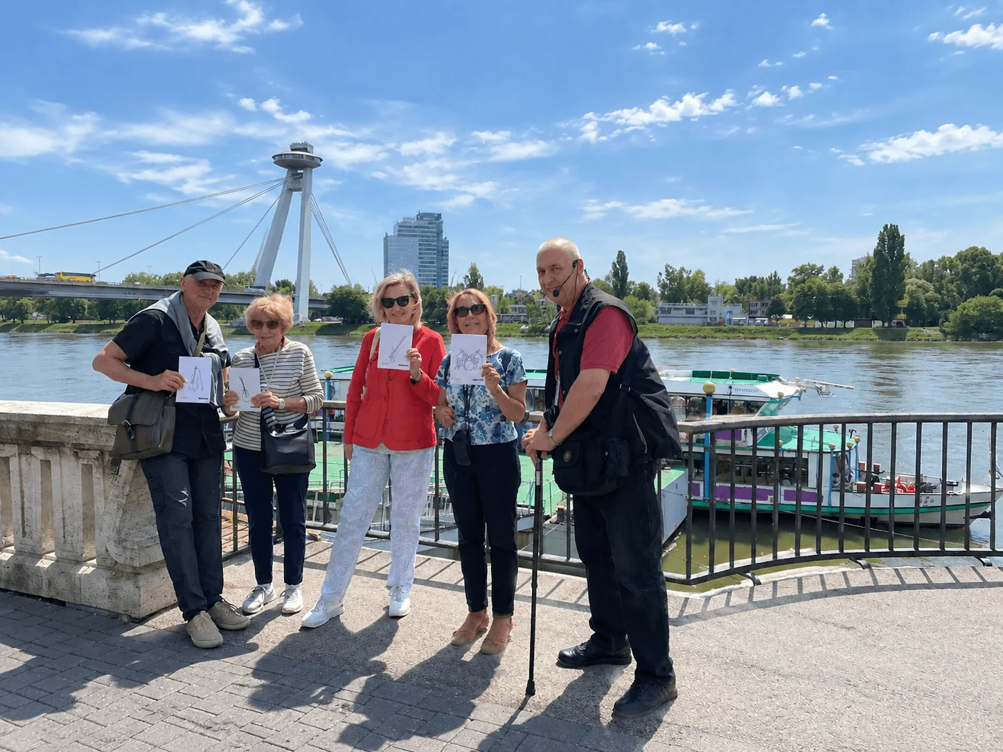 Un guide local avec un petit groupe sur le quai du Danube à Bratislava, avec le Most SNP (le pont UFO) et un bateau fluvial amarré derrière eux.