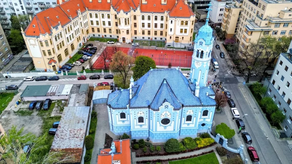 An aerial view of the Blue Church of St Elizabeth in Bratislava, its pale blue walls, tiled roof and round tower standing out among the surrounding streets.