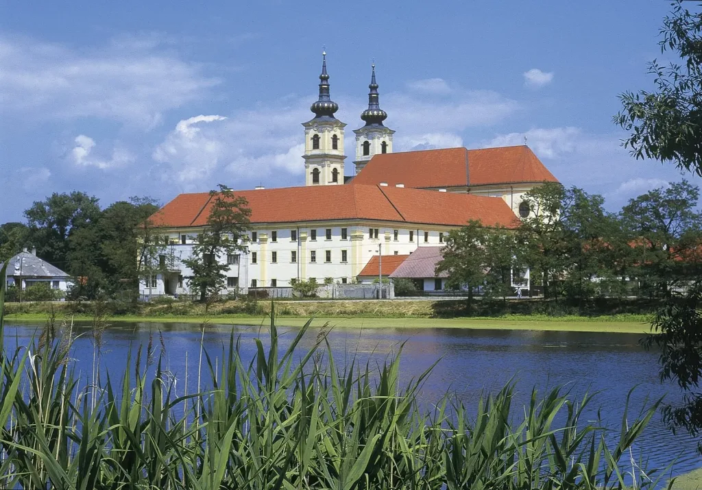 The twin-towered national basilica at Šaštín with its long red roof and monastery wings, seen across a reed-fringed lake.
