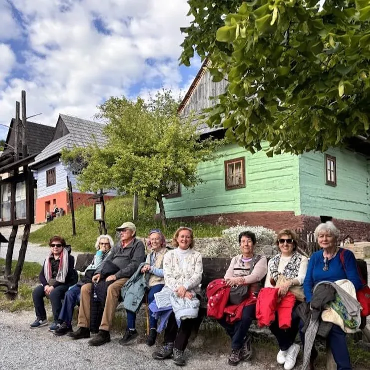 A tour group sitting on a bench in front of painted wooden log houses in a Slovak village.